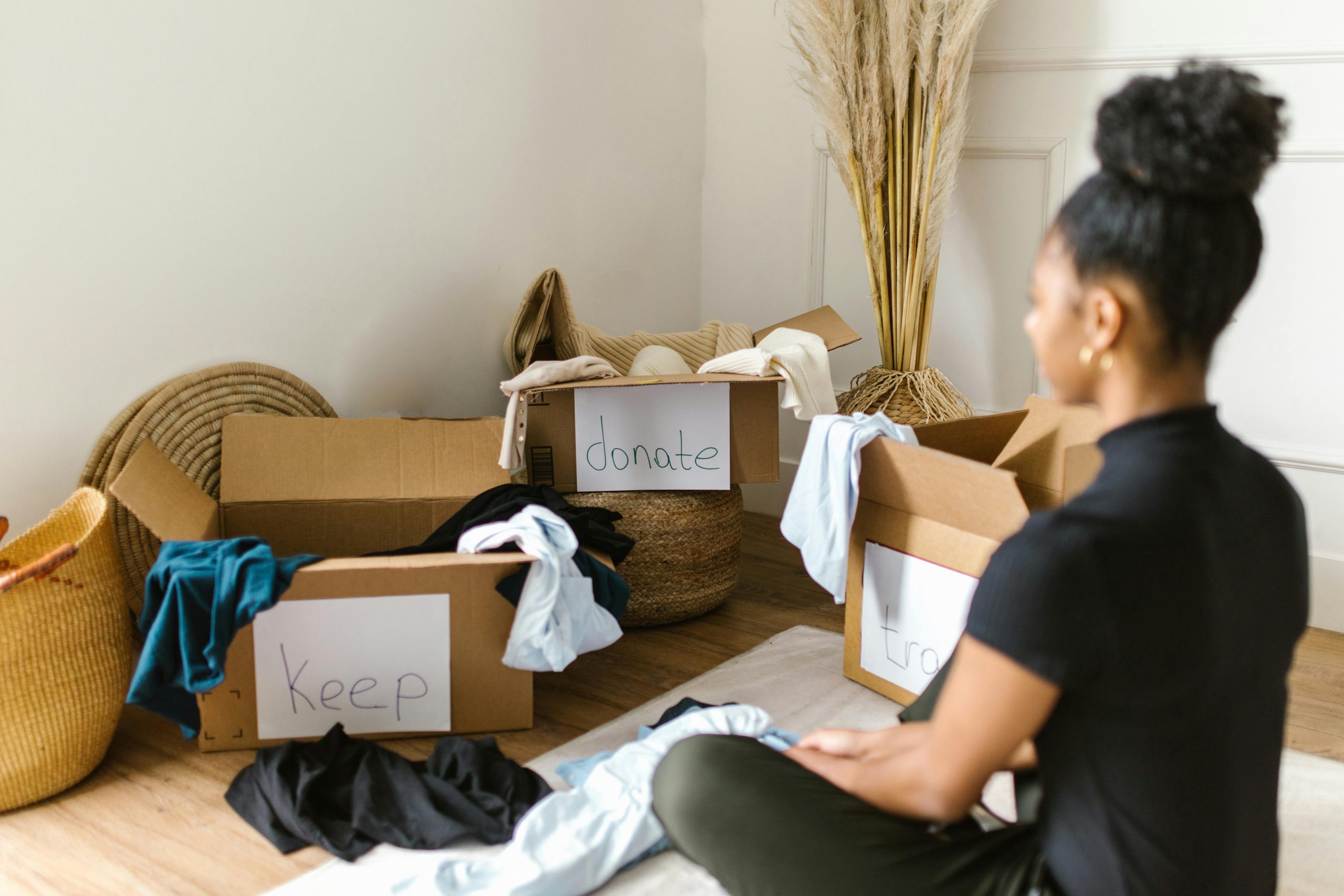 woman sitting on the floor, with clothes in cardboard boxes labeled 'keep,' 'donate,' and 'trash'